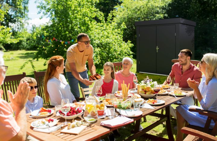 Man erkennt eine fröhliche Familie, die gemütlich in ihrem Garten grillt.