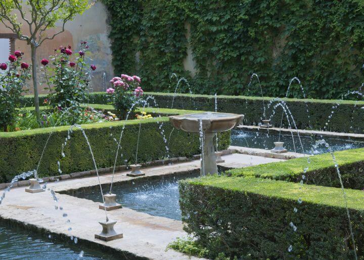 Springbrunnen neben Weg, rundherum Hecken und Rosen