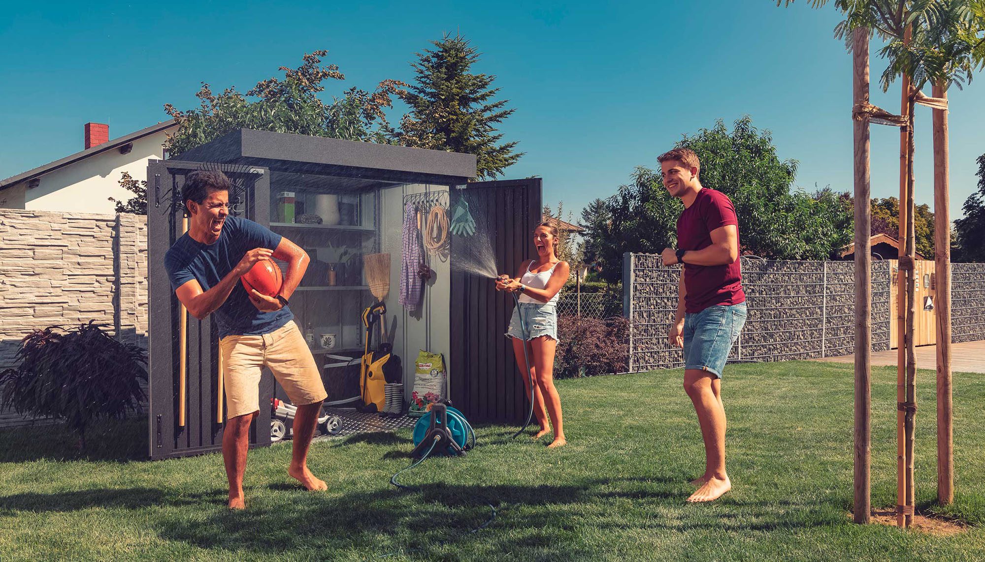 Ilesto Gartenhaus in anthrazit steht geöffnet im Garten, darin sind einige Gartenutensilien zu sehen und davor spielt eine junge Gruppe von Menschen im Garten