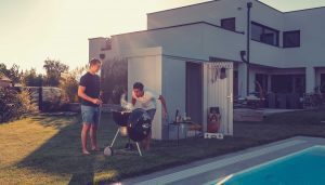 Ilesto Gartenhaus in silber steht geöffnet am Rand eines Pools, davor stehen zwei junge Männer am grillen