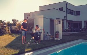 2 men having a bbq/ silver garden shed behind them / white house in BG