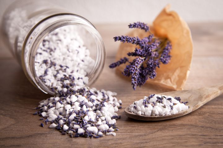 Lavendel Badesalz läuft aus einem liegenden Glas heraus, daneben liegt ein Strauch violetter Lavendelblüten