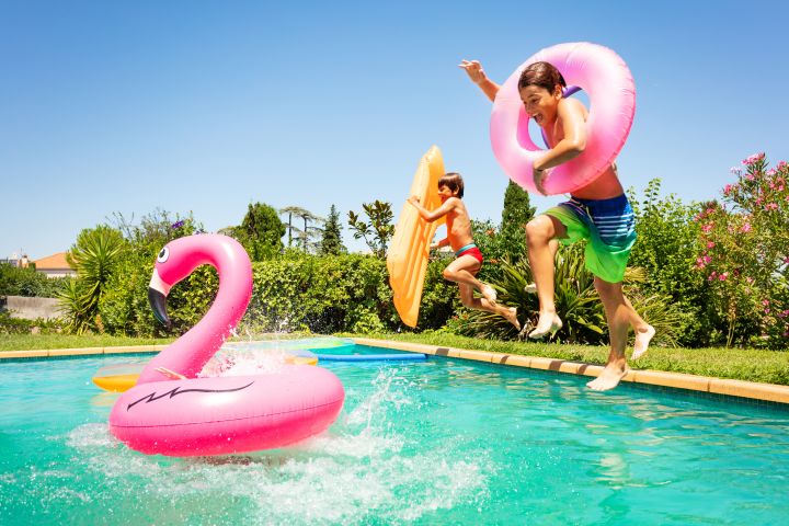 Kinder springen in einen Pool, einer hält einen Schwimmreifen, der andere eine Luftmatratze in der Hand