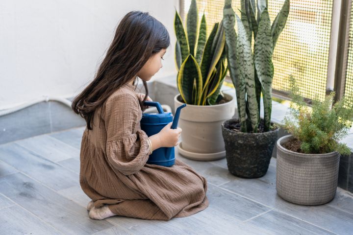 Kleines Mädchen in braunem Kleid wässert Pflanzen auf dem Balkon