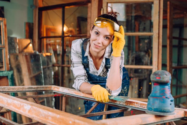 Nahaufnahme einer Frau, die gelbe Arbeitshandschuhe und eine Schutzbrille trägt und direkt in die Kamera sieht. Sie schleift einen Tisch mit Sandpapier ab. Upcycling Konzept.